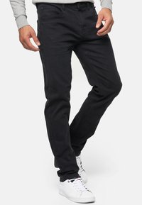 Man die zwarte slim-fit jeans en witte sneakers draagt, met gekruiste benen tegen een effen achtergrond staat.