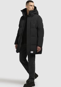 Mustankate pufferskoht, põlvpikkusega, lukuga ja nööbiga kinnitamiseks, soonikkoes sisemise kaeluse ja väliste lukuga taskutega, kantud musta riietuse peal, kombineeritud tumedate teksadega.