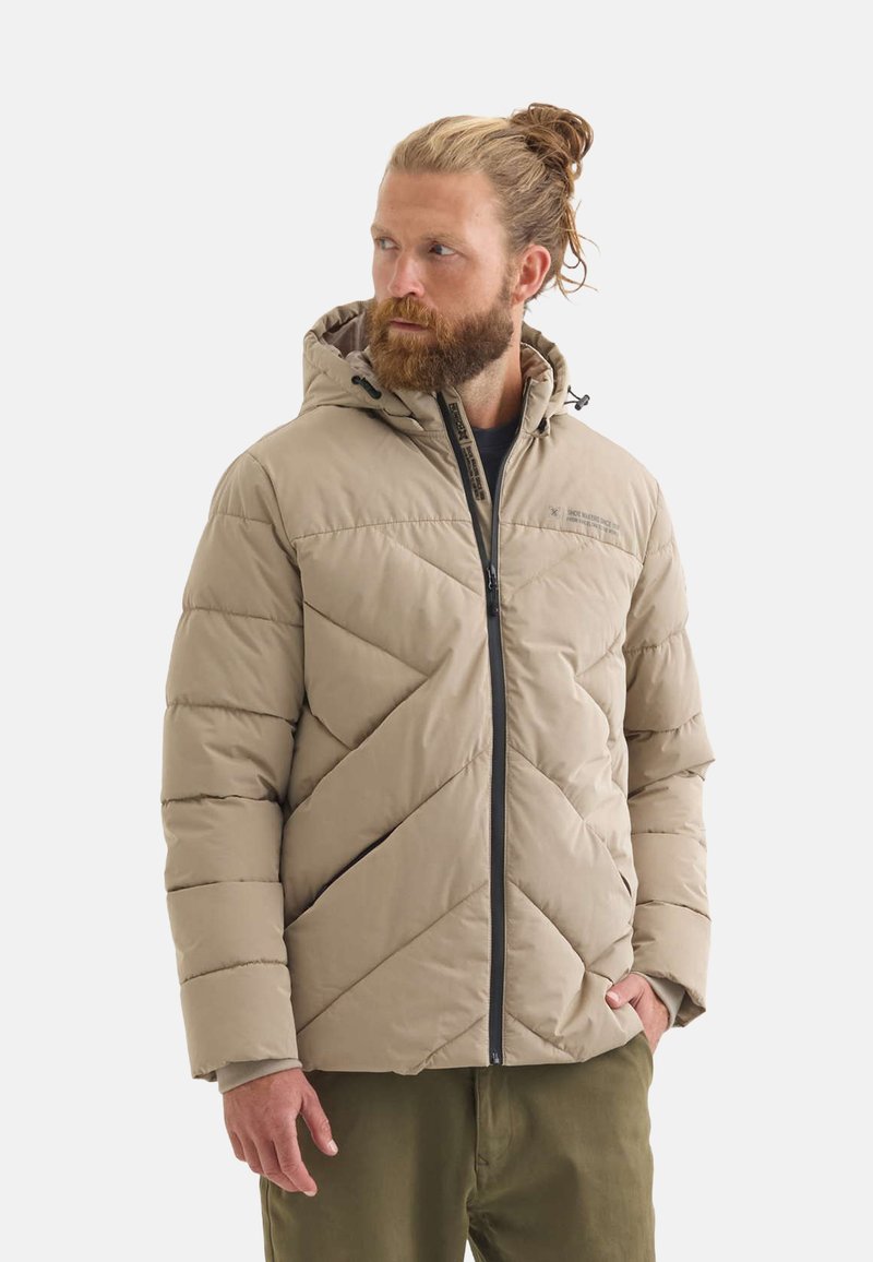 Chaqueta acolchada beige con patrón de acolchado diagonal, cierre de cremallera, capucha y detalles en negro. Lleva puesta sobre una camiseta oscura y pantalones verde olivo.