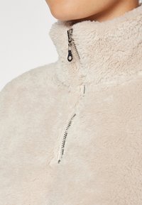 Fleece zippé beige clair avec col haut, tissu texturé doux, et une fermeture éclair proéminente avec des accents métalliques.