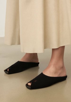 Lábak fekete, nyitott orrú papucsban, hosszú bézs szoknyával, sima, világosbarna padlón állva.