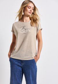 Camiseta de manga corta beige con un corte relajado, con el texto "Bon" impreso en negro y gris. Llevada con pantalones de pierna ancha azules.