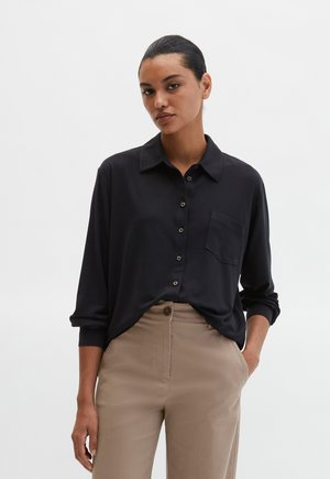 Chemise noire à boutons avec une poche, manches longues et coupe décontractée, associée à un pantalon beige avec fermeture à boutons sur le devant.