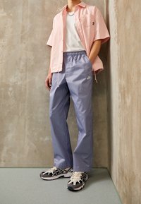 Camicetta rosa a maniche corte, t-shirt bianca sotto, pantaloni lilla chiaro con vita elastica e scarpe da corsa nere e beige. Sfondo in cemento.