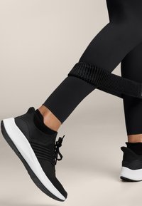 Fekete leggingsben lévő lábak, egy ellenállás szalag egy comb körül, fekete és fehér sportcipőt viselve, edzéshelyzetben.