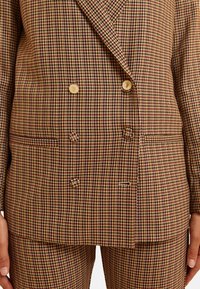 Blazer de doble botonadura en un patrón de cuadros de lana marrón y amarillo. Presenta botones dorados, tejido texturizado y dos bolsillos frontales.