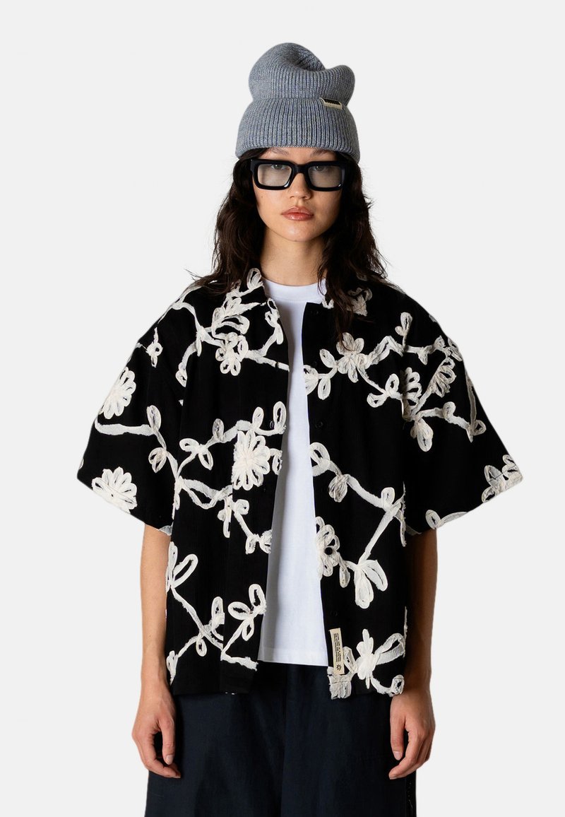 Personne portant une chemise surdimensionnée à fleurs noires et blanches, un sous-maillot blanc, un bonnet en tricot gris et de grandes lunettes carrées noires, faisant face à l'avant.