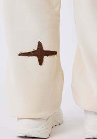 Crèmekleurige sweatpants met een bruine gestikte kruisaccent op het onderbeen, gecombineerd met witte sneakers. Zacht, casual materiaal.