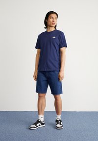 Navy zils kokvilnas T-krekls ar baltu Nike logo, apvienots ar džinsu šortiem un melnbaltiem sporta apaviem. Fona teksturēta grīda.