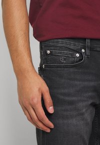 Czarne dżinsy z denimu o wąskim kroju, zawiązane srebrnymi elementami, subtelnie wyblakłe, z wyszywanym logo na przedniej kieszeni. Ręka oparta na dżinsach.