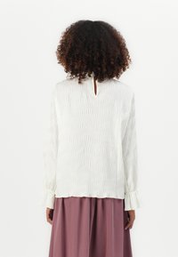 Donna con capelli ricci che indossa una blusa bianca testurizzata con polsini arricciati e pantaloni larghi color malva, in piedi di spalle.