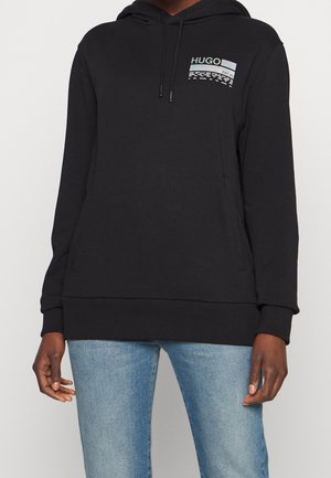 Schwarzer Hoodie mit Fronttasche und "HUGO"-Logo auf der linken Brust, getragen mit hellblauen Jeans von einer Person mit dunklem Hautton.