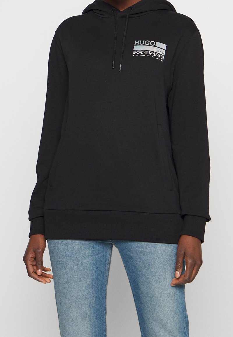 Pull à capuche noir avec poche avant et logo "HUGO" sur le côté gauche de la poitrine, porté avec un jean bleu clair sur une personne à la peau foncée.