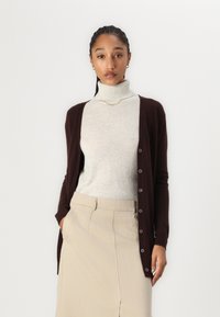 Hnědý cardigan přes světle šedý rolák, doplněný béžovou sukní. Cardigan má knoflíky a jemnou texturu. Jednoduchý, elegantní design.