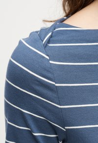 Nahaufnahme von Schulter und Hals einer Person, die ein blaues Hemd mit weißen horizontalen Streifen trägt.