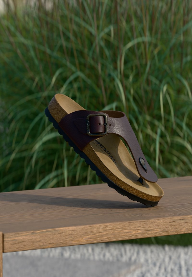 Καφέ δερμάτινο σανδάλι Birkenstock με πόρπη και πάτο από φελλό, ισορροπεί σε ξύλινη επιφάνεια, φόντο με πράσινο γρασίδι.