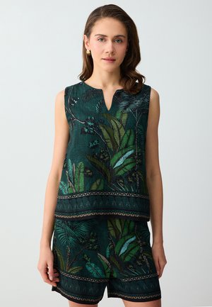 Femme portant un haut sans manches vert foncé assorti et un short avec des motifs de feuilles et de branches tropicales sur un fond uni.