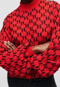 Red sweater with black K logo pattern, ribbed collar and cuffs, textured fabric, and relaxed fit. Styled with crossed arms.