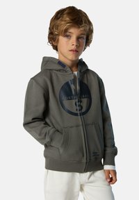 Junge mit lockigem blonden Haar trägt einen grauen Zip-Hoodie mit großem, kreisförmigem North Sails-Logo und weißen Hosen und steht vor einem schlichten Hintergrund.