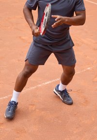 Tennisimängija hallis sportlikus särgis ja lühikeses pantaloonis seisab saviväljakul, hoides punast ja musta reketti, jalas tumesinised tennisetossud valgete sokkidega.
