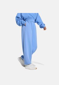 Persona vistiendo pantalones de chándal sueltos de color azul claro y sudadera a juego con zapatillas blancas, con una mano en el bolsillo, caminando sobre un fondo liso.