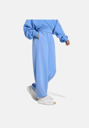 Persoon in losse lichtblauwe trainingbroek en bijpassende sweatshirt met witte sneakers, hand in zak, wandelend op een effen achtergrond.
