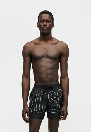 Homme athlétique à la peau foncée debout, portant un short de bain noir avec de grandes lettres blanches "MOSC" à l'avant.