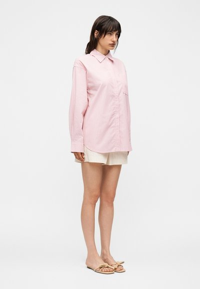 Γυναίκα με σκουρόχρωμα μαλλιά, που φοράει ένα φαρδύ ροζ ριγέ μακρυμάνικο πουκάμισο, μπεζ σορτς και μπεζ σανδάλια με φιόγκους, στέκεται μπροστά σε απλό λευκό φόντο.