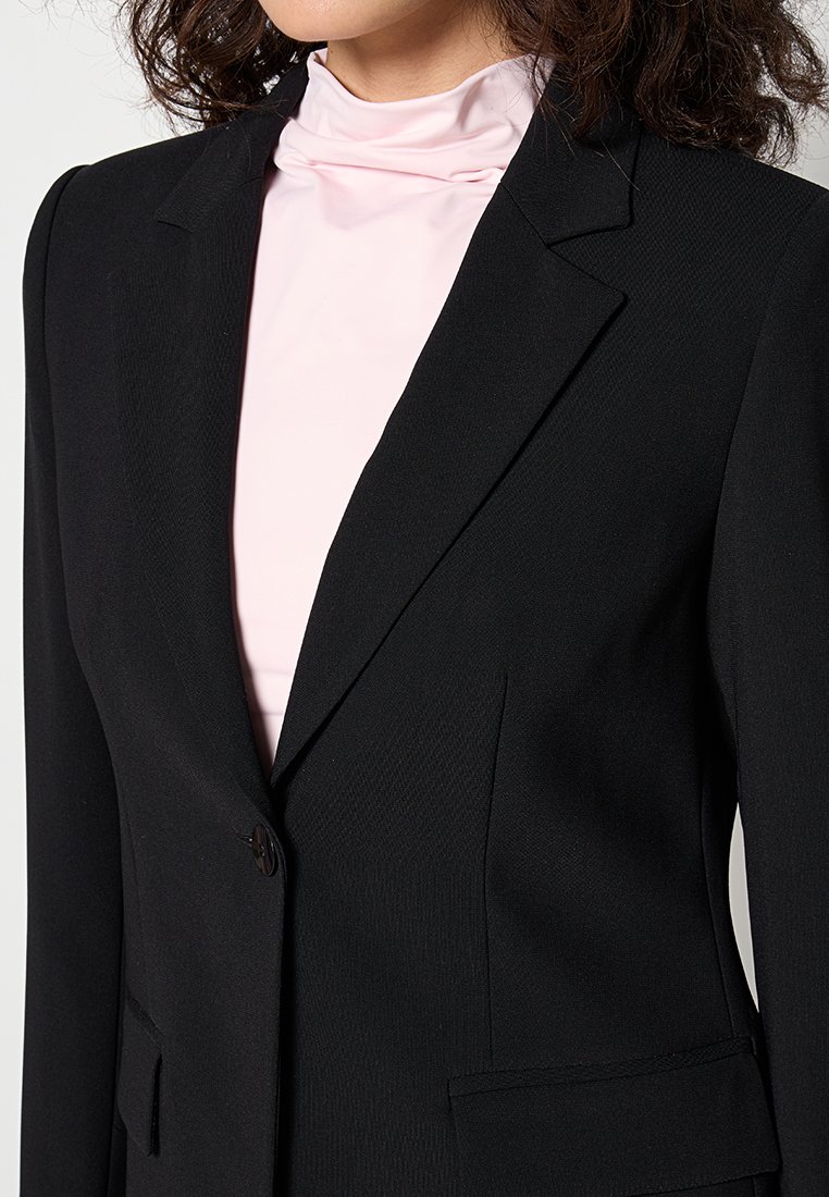 Blazer noir ajusté avec une finition texturée, fermeture à un bouton et revers pointus, superposé à un haut rose clair à col montant.
