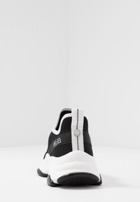 Chaussure de sport noire avec une tige en mesh, des accents blancs, un talon texturé et une languette de tirage. Elle possède une semelle intermédiaire blanche épaisse et une semelle extérieure en caoutchouc noir.