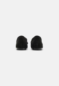 Mocassins pretos em camurça com uma textura suave, bico redondo, perfil baixo e um discreto detalhe de costura ao longo das bordas.