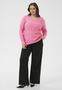 Ružový sveter s výstrihom do V z jemného, texturovaného materiálu, kombinovaný s čiernymi širokými nohavicami a čiernymi lodičkami s špicatou špičkou na neutrálnom pozadí.