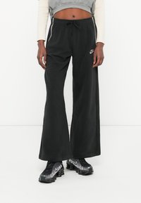 Črne široke trenirke iz mehkega materiala, ki imajo oblačilni pas na vrvico in majhen bel Nike logo na levi strani.
