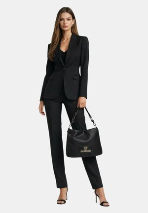 Donna in completo nero su misura e tacchi che tiene una grande borsa nera Love Moschino su sfondo uniforme.