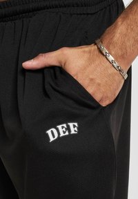 Sorte atletiske joggers med en struktureret overflade, der har et hvidt "DEF" logo over sidelommen og en elastisk talje.