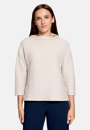 Femme aux longs cheveux bruns portant un haut à manches longues clair et texturé ainsi qu’un pantalon sombre, debout devant un fond uni.