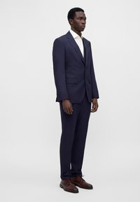 Homme debout portant un costume bleu foncé avec une chemise blanche et des chaussures habillées en cuir marron sur un fond clair uni.