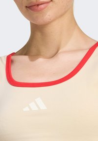 Top sportivo beige con collo rotondo rosso e texture sottile, con il logo bianco Adidas sul petto.