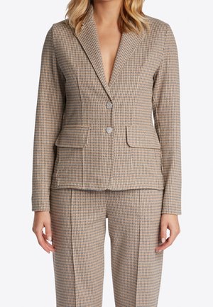 Primer plano de una persona que lleva una chaqueta con estampado pata de gallo en beige y azul y pantalones a juego con dos bolsillos delanteros y botones metálicos.