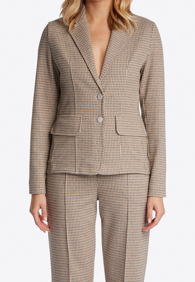 Gros plan d'une personne portant un blazer à motif pied-de-poule beige et bleu et un pantalon assorti avec deux poches avant et des boutons métalliques.