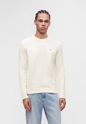 Ung mand med flettet hår iført en cremefarvet sweater og lyseblå jeans, stående mod en ensfarvet grå baggrund.