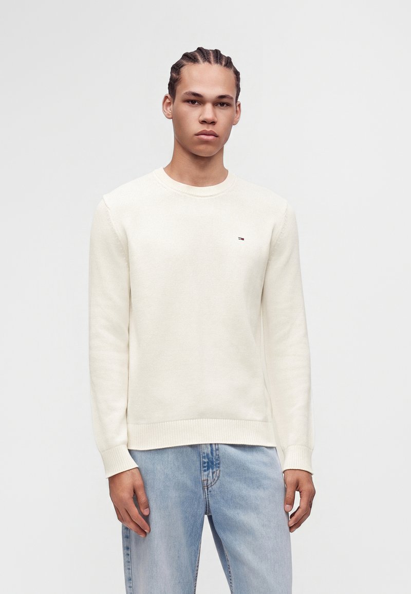 Jeune homme aux cheveux tressés portant un pull crème et un jean bleu clair, debout devant un fond gris uni.