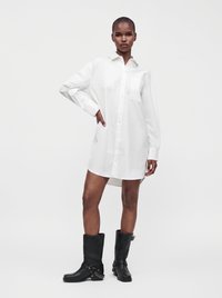 Jeune femme portant une robe chemise blanche boutonnée et des bottes noires mi-mollet avec détail chaîne, debout avec une main sur la hanche.