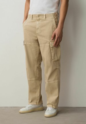 Beige Cargohose mit Seitentaschen, getragen von einer Person in weißen Sneakers und weißem T-Shirt, stehend vor einer schlichten hellen Wand.