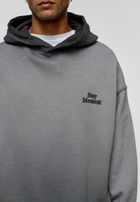 Grijze hoodie met een kwetsige stof, met een zwarte geborduurde "Stay Moment" detail aan de voorkant.