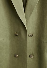 Olive green blazer with a textured fabric, double-breasted design, and two large brown buttons. Features side pockets and a tailored fit.