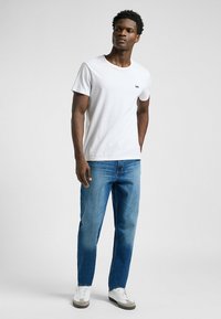 Camiseta blanca de manga corta con un pequeño logo negro de "Lee", combinada con jeans rectos de un azul medio y zapatillas blancas.