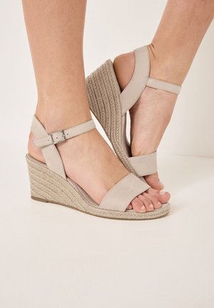 Beige kilehæle sandaler med ankelremme, tekstureret rebdetalje på kilen, åben tå-design, båret med en denimnederdel på et trægulv.