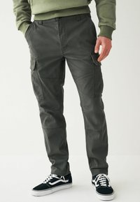 Cargo-Hosen aus dunkelgrauem Stoff mit schmaler Passform. Verfügt über mehrere Taschen und gestickte Akzente. Kombiniert mit schwarzen Sneakers.
