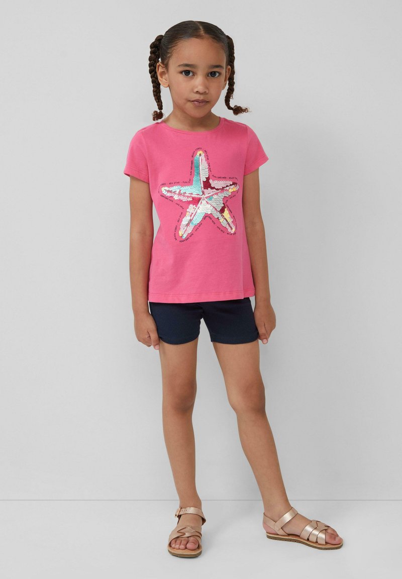 Roze katoenen T-shirt met een sterontwerp van pailletten, gedragen met marineblauwe shorts en gouden sandalen. Eenvoudige ronde hals en korte mouwen.